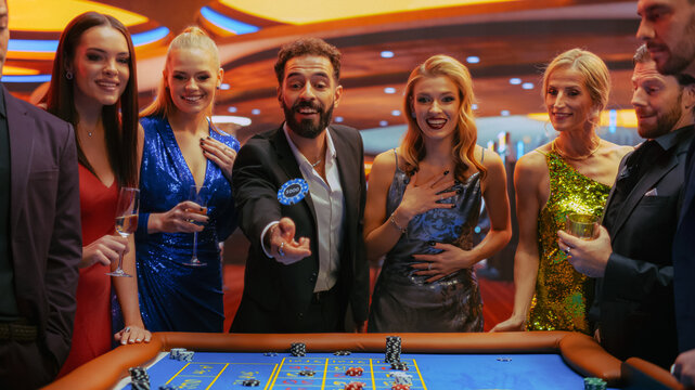 Cheerful Group of Rich Young People Gathered Around a Roulette Table at a Modern Casino. Handsome Hispanic Male Throwing a 5000 Dollar Casino Coin Towards the Camera, Crowd Cheering, Having Fun.