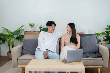A young Asian couple sits comfortably on a beige couch, engaging in a pleasant conversation in a well-lit living room.