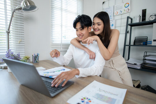 A smiling young Asian couple collaborates on a project, works together managing their budget, working on a laptop in a home office setting.