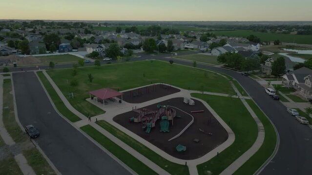 This video shows an aerial view of a neighborhood park with slides, monkey bars, swings, picnic tables with a shelter, and a green grass field. It was taken on a partly cloudy summer morning.