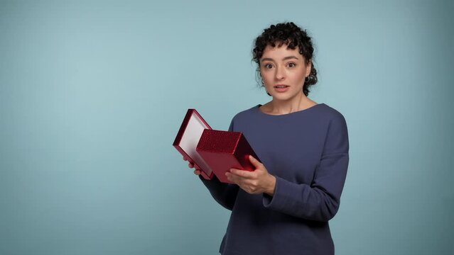 Shocked curly woman opens present red box surprised looks camera and says wow. Excited positive female wearing blue sweater with gift in hands on isolated light blue background with copy space