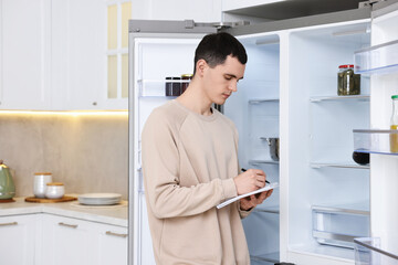 Man writing notes near empty refrigerator in kitchen