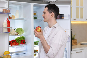 Happy man holding apple near refrigerator in kitchen