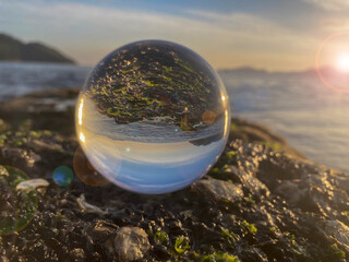 Glass ball on the sea rock with crustaceans and algae reflecting the natural landscape of the sea during sunset. In the background the sea, horizon and islands in the bay. Sun reflections in the lens.