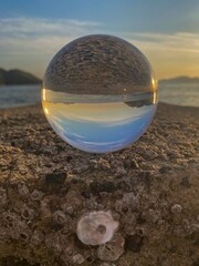 Glass ball on the sea rock with crustaceans reflecting the natural landscape of the sea during sunset. In the background the sea, horizon and islands in the bay. Sun reflections in the lens.