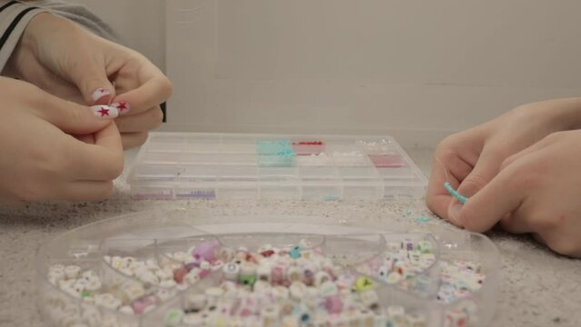 close-up of hands of brother and sister seated on carpet, threading beads onto string to make bead bracelets and jewelry. Concept of children's activities at home and development of fine motor skills