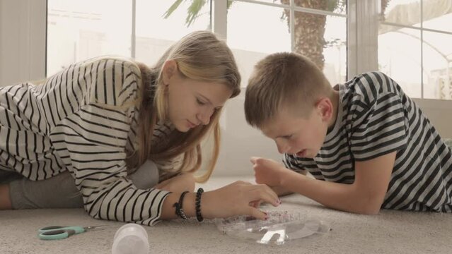 Generation Z teenagers, boy and a girl, sit on floor in bright living room, weaving bracelets from beads, helping each other out
