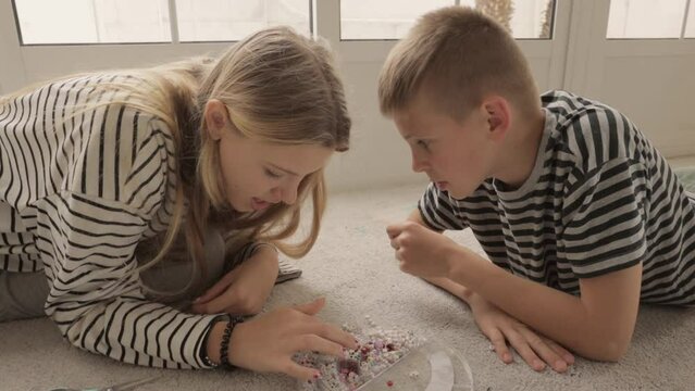 brother and sister lie on  carpet in bright living room with large windows, making bracelets from beads, joyfully chatting with each other