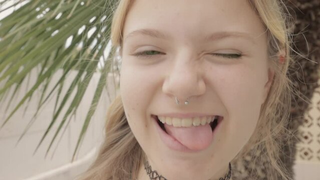 teenage hipster girl from Generation Z with septum piercing in her nose, against  backdrop of palm trees, turning towards camera with smile and sticking out her tongue