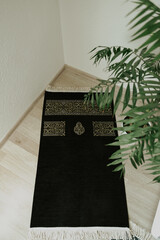 Black velvet prayer mat with a Kaaba pattern in the corner against a background of a house plant