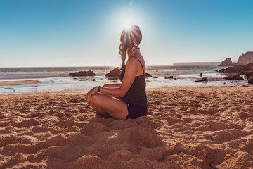 Ragazza seduta a gambe incrociare sulla riva del mare con il sole contro luce - obrazy, fototapety, plakaty