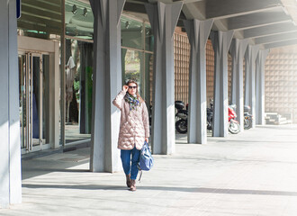 Beautiful stylish young woman with bag, sunglasses and mobile phone standing near building on the street