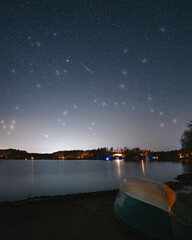 A night sky view above a pond with a rowboat on the shore of a sandy beach and homes along the...