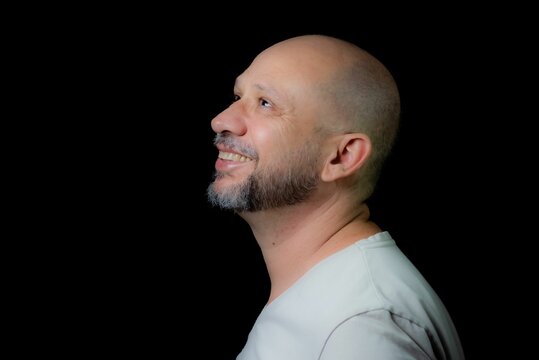 Portrait of a bald bearded man looking up and smiling against a black background