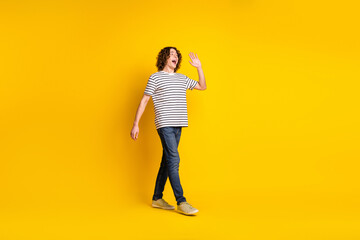 Full size portrait of nice young man walk empty space arm wave hi wear striped t-shirt isolated on yellow color background