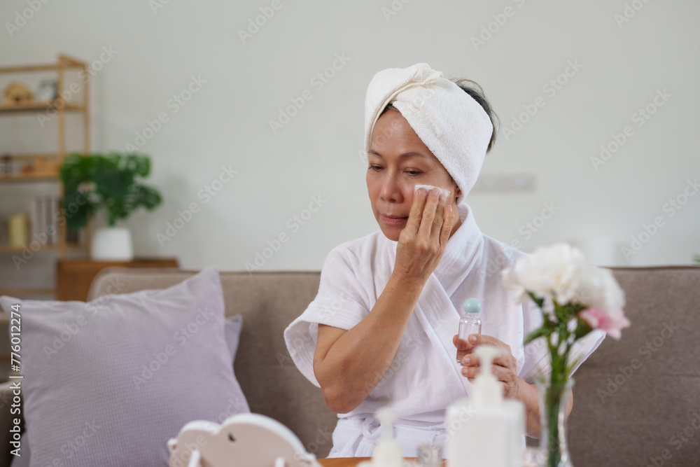 Wall mural mature woman in a self-care routine, gently applying facial toner, sitting comfortably in her living