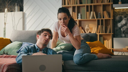 Couple making purchase online looking at laptop sitting couch cozy living room.
