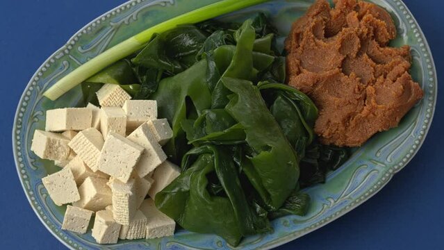 Ingredients for vegan miso soup: miso paste, rehydrated wakame seaweed, cubed tofu, scallion. Blue background. Table spin.