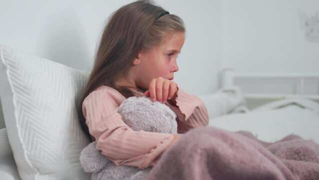 Upset sick girl coughing while sitting with soft toy on bed at home. Sad girl child suffering from cold and flu and using digital tablet in apartment.