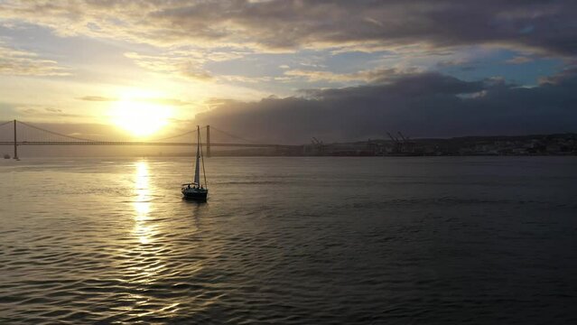 25 April Suspension Bridge at Sunset and Silhouette of Sailing Yacht. Ponte 25 de Abril and Tagus River. Portugal. Aerial View. Golden Hour. Orbiting