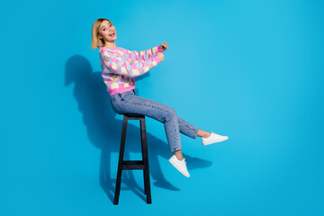 Full length side profile photo of ecstatic girl wear pullover sit on chair hold invisible steering wheel isolated on blue color background