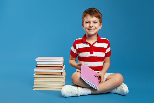 Little schoolboy in red striped shirt, holding book in hands and looking at camera while sitting on studio floor near bunch of books, isolated over blue background. Back to school concept - Powered by Adobe