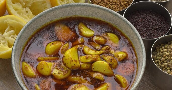 Garlic pickle, side dish from India. Around the bowl with pickled garlic are containers with spices used for pickling and squeezed lemons. Table spin.