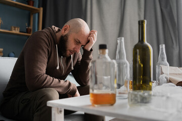Alcohol addiction abuse and alcoholism. Upset millennial man drinker alcoholic sitting at home with empty bottles drinking whiskey alone, sad depressed addicted man having problem.