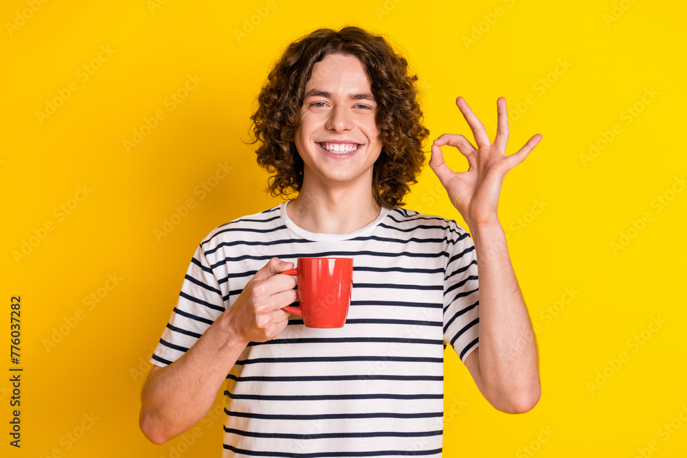 Poster photo of nice young man hold mug show okey symbol wear striped t-shirt isolated on yellow color back