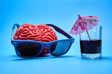 Top view of a red plastic human brain wearing blue sunglasses next to a cocktail party with an umbrella on a blue background.