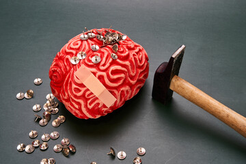 Red human brain on top of a pile of thumbtacks, with a black hammer next to it on a dark background. Representation of Headache