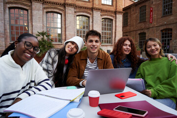 Multiethnic group of student friends gathering at university campus to study all together for a school project, sitting outside in a table using a laptop, looking at camera smiling.