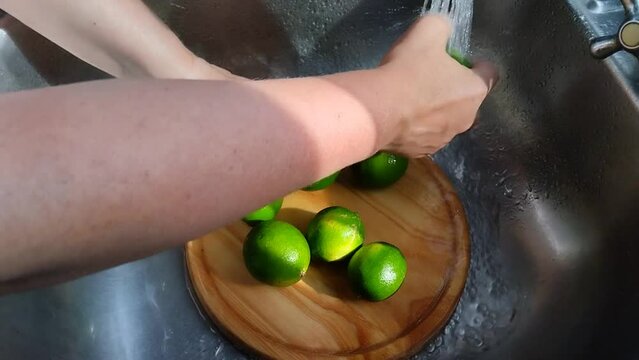 Unas manos de mujer lavando  varios limones verdes con agua, colocados en una tabla de madera en un fregadero