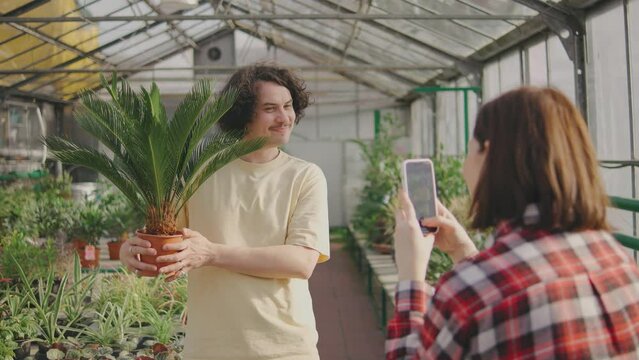Woman photographing a man with a plant for his blog, Man and woman having fun together. Couple chose a beautiful green plant and take a pictures with it on a smartphone and are happy. Funny couple.