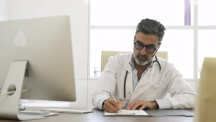 Male doctor noting something on document and using computer in clinic