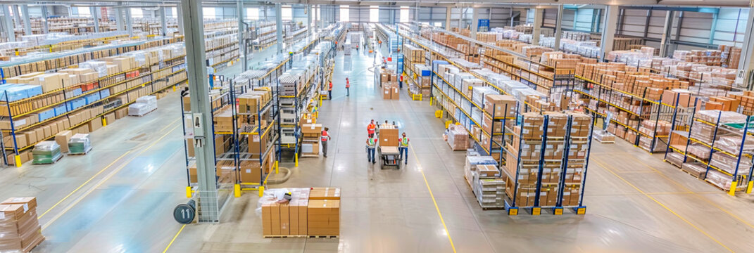 Warenlager mit gut organisierten Regalen und Stapeln von Kartons und Paletten. Mitarbeiter sind dabei, Waren zu kommissionieren, zu verpacken und f&uuml;r den Versand vorzubereiten. Gabelstapler und andere