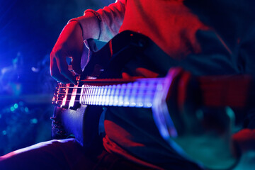Cropped photo of guitarist's fingers on fretboard of electric guitar with vibrant blue-red stage...
