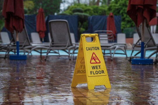 Yellow warning sign, stating "Caution Wet Floor", next to a hotel resort's swimming pool and sun loungers.