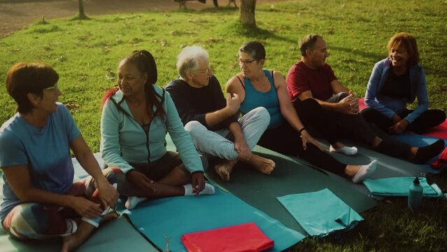 Group of multiracial senior friends taking a break sitting on mat after workout activity - Health elderly people lifestyle