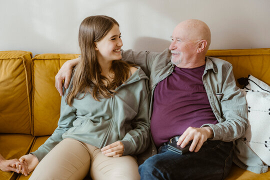 Smiling senior man sitting with arm around teenage granddaughter on sofa at home