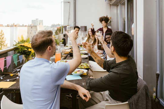 Men toasting drinks with group of friends sitting at dining table in balcony