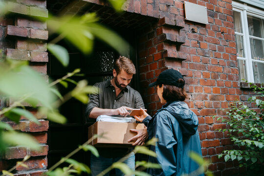 Mature man doing electronic signature while receiving parcel from female delivery person standing at doorstep