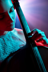 Close up portrait of female cellist performing classical melodies with radiant purple stage lights behind her on stage. Concept of hobby and work, music festivals, concerts, symphony show, culture.