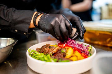 A chef in black gloves prepares a vibrant salad with grilled meat, fresh greens, and colorful...