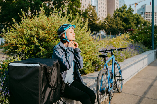 Female delivery person fastening helmet while sitting on bench by bicycle at footpath