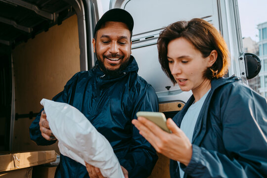 Smiling male delivery person showing parcel to female coworker holding smart phone near van