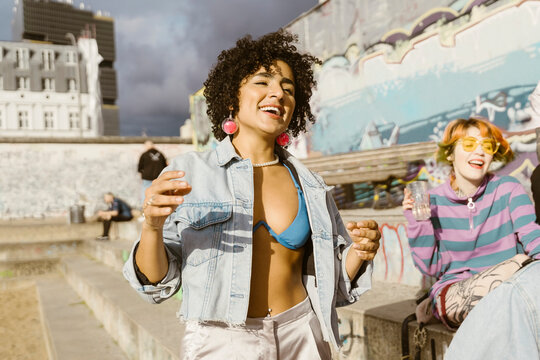 Carefree smiling non-binary person dancing with friends sitting on steps in city at sunny day