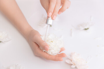 Naklejka premium Organic oil for health of nail and skincare concept. Women hands hold pipette with cosmetic serum on white background with flowers, top view