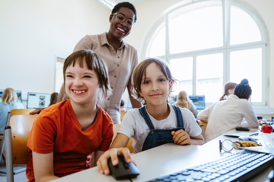 Portrait of smiling girl next to female friend with disability sitting in computer class at school