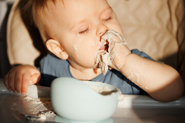Happy messy baby boy enjoys food, sits In high chair and eats porridge with his hands without...